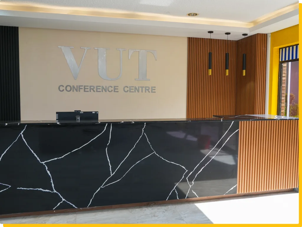 Reception area with a beige wall displaying 'VUT CONFERENCE CENTRE', a black marble counter, and a vertical wooden slat wall behind it.