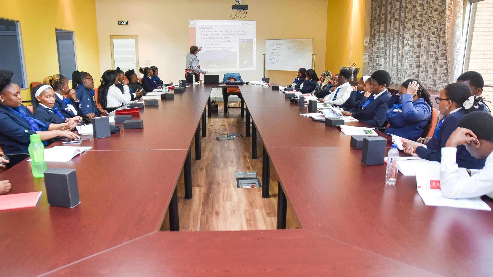 A classroom-style conference room with students seated around a U-shaped table, listening to a lecturer at the front presenting slides on a screen.