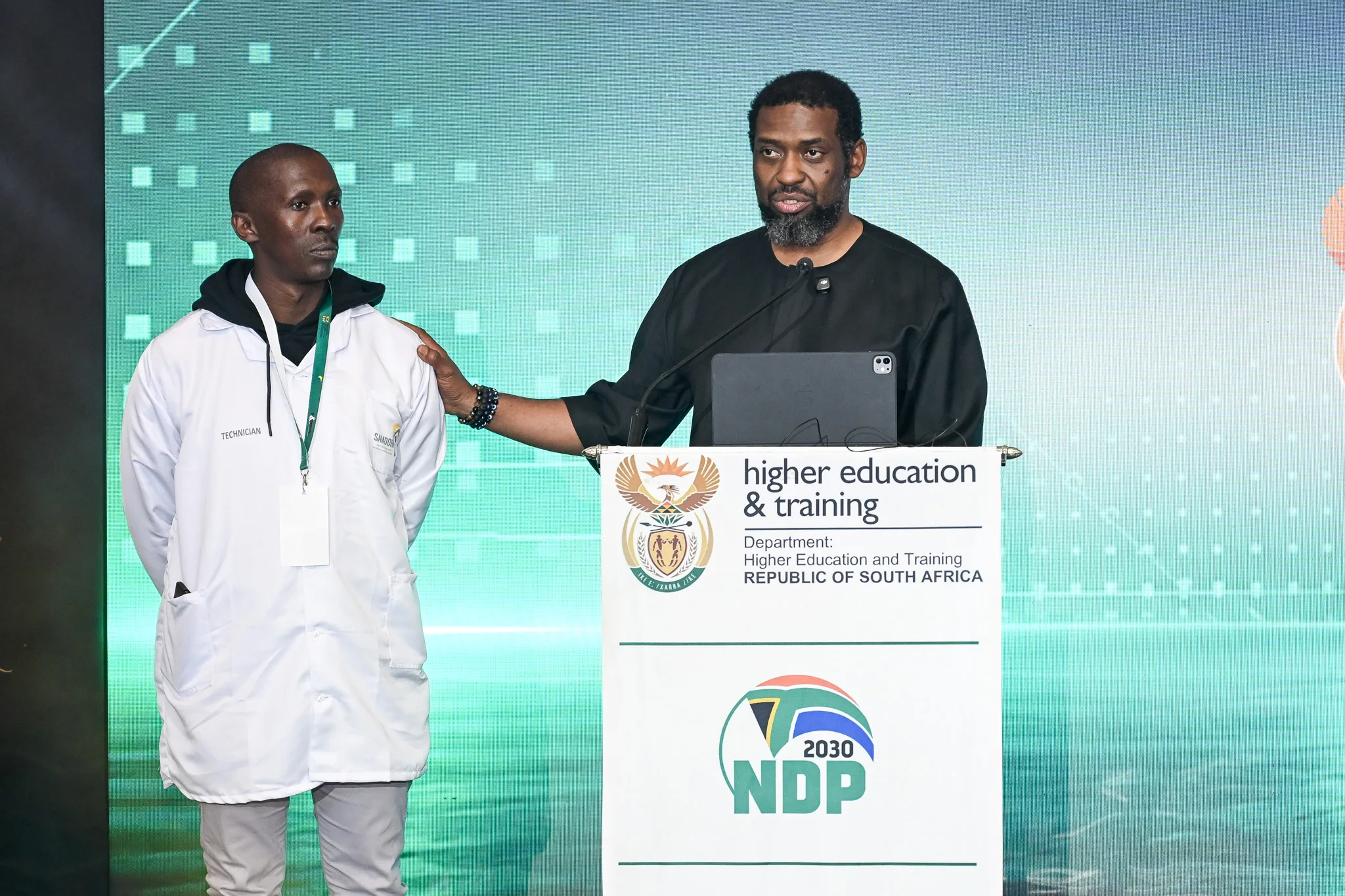 Minister of Higher Education and Training, Buti Manamela, stands on stage with one of the 25 young entrepreneurs who received cellphone repair containers.