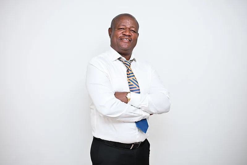 Smiling man in a white shirt and striped tie poses with arms crossed against a light background, exuding confidence and approachability.