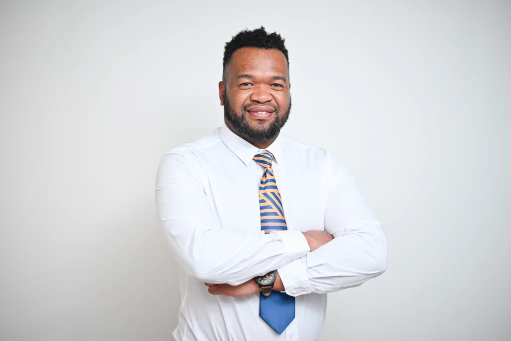 Professional headshot of a smiling man in a white shirt and striped blue-orange tie with arms crossed against a neutral background.