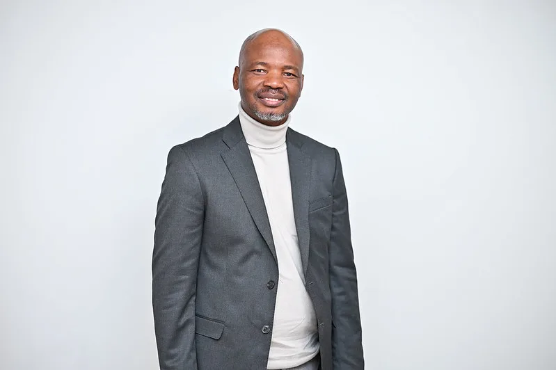 Smiling man in a gray suit and white turtleneck posing against a plain light background.