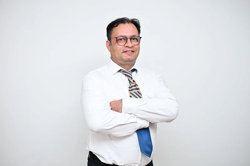 Professional man posing with arms crossed in a white shirt and patterned tie against a light background, wearing glasses.