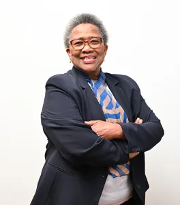 Portrait of a smiling, senior Black woman with short gray hair and red glasses, wearing a navy blazer and blue striped scarf, arms crossed.