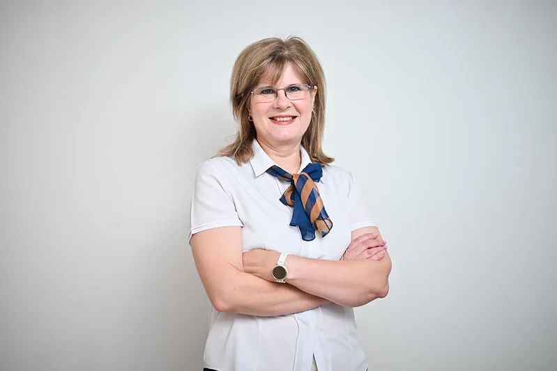 Professional woman with arms crossed, smiling, in a white blouse and blue scarf against a light background.