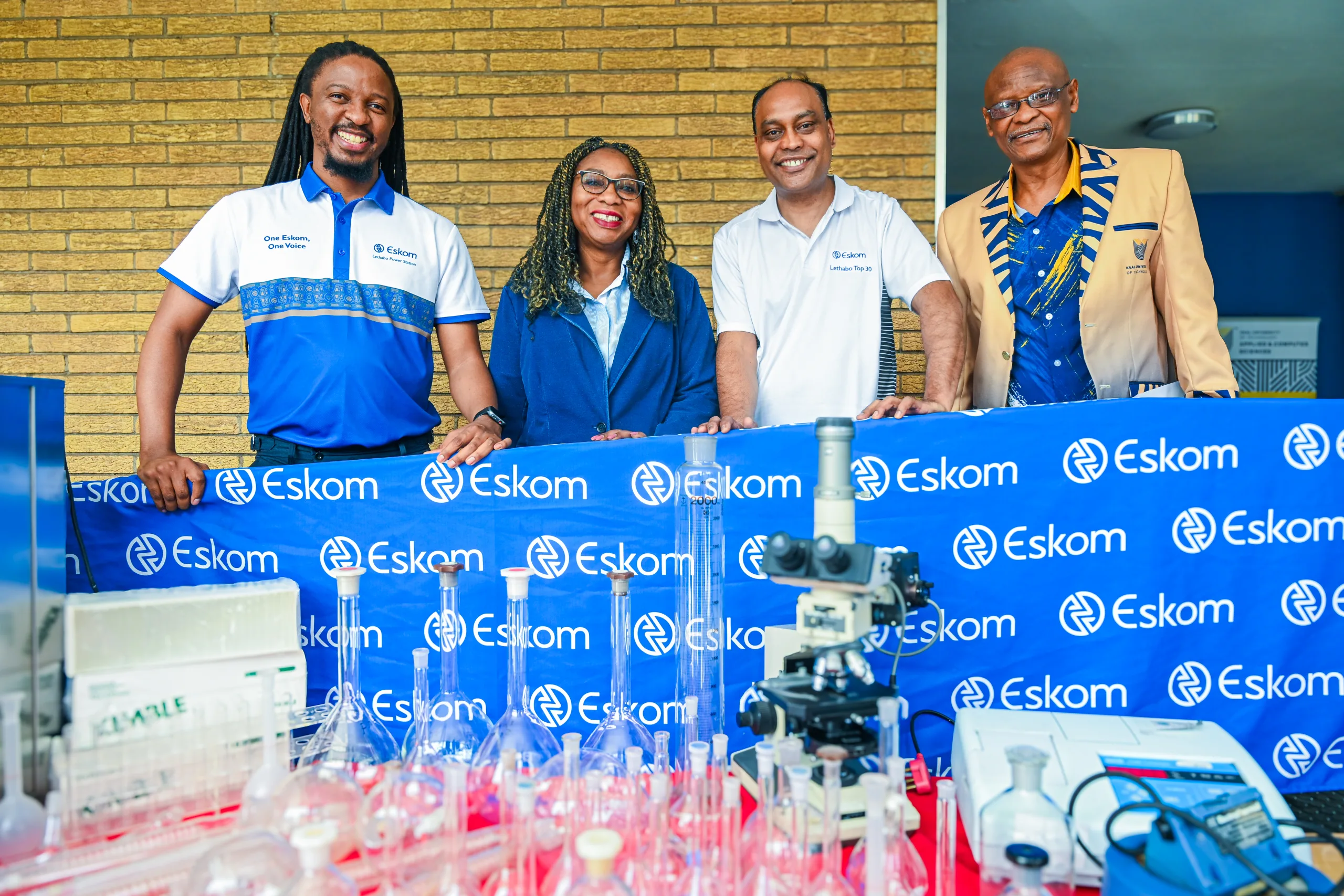 Four professionals smile for a group photo behind a table of lab glassware at an Eskom-branded event.