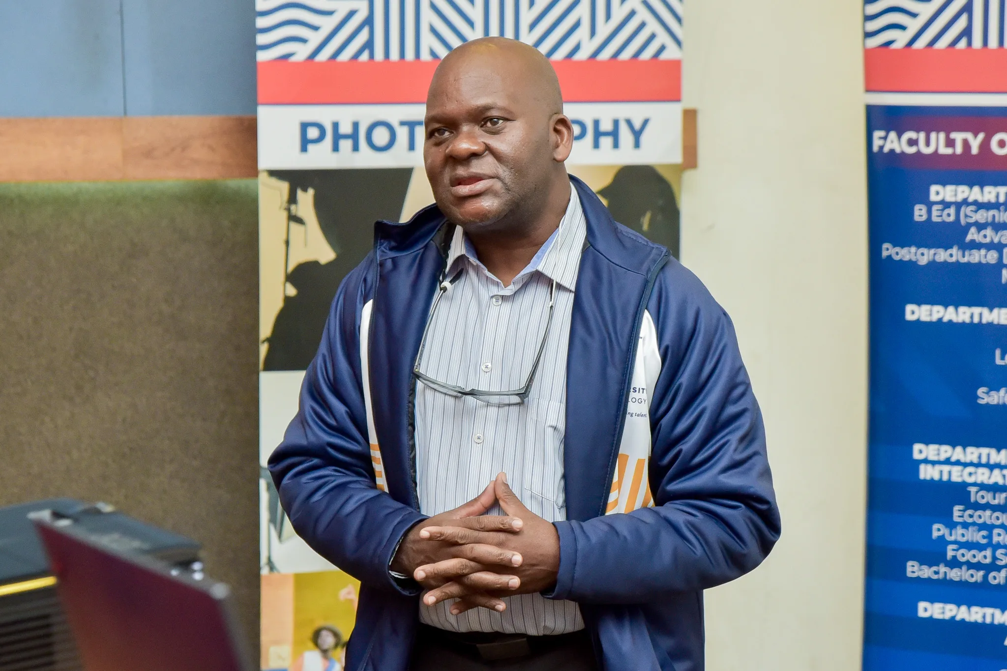 FHS Postgraduate colloquium-19 – Vaal University of Technology – Vaal University of Technology Man in a blue jacket speaks at a campus event, hands clasped, with 'PHOTOGRAPHY' and department banners in the background.