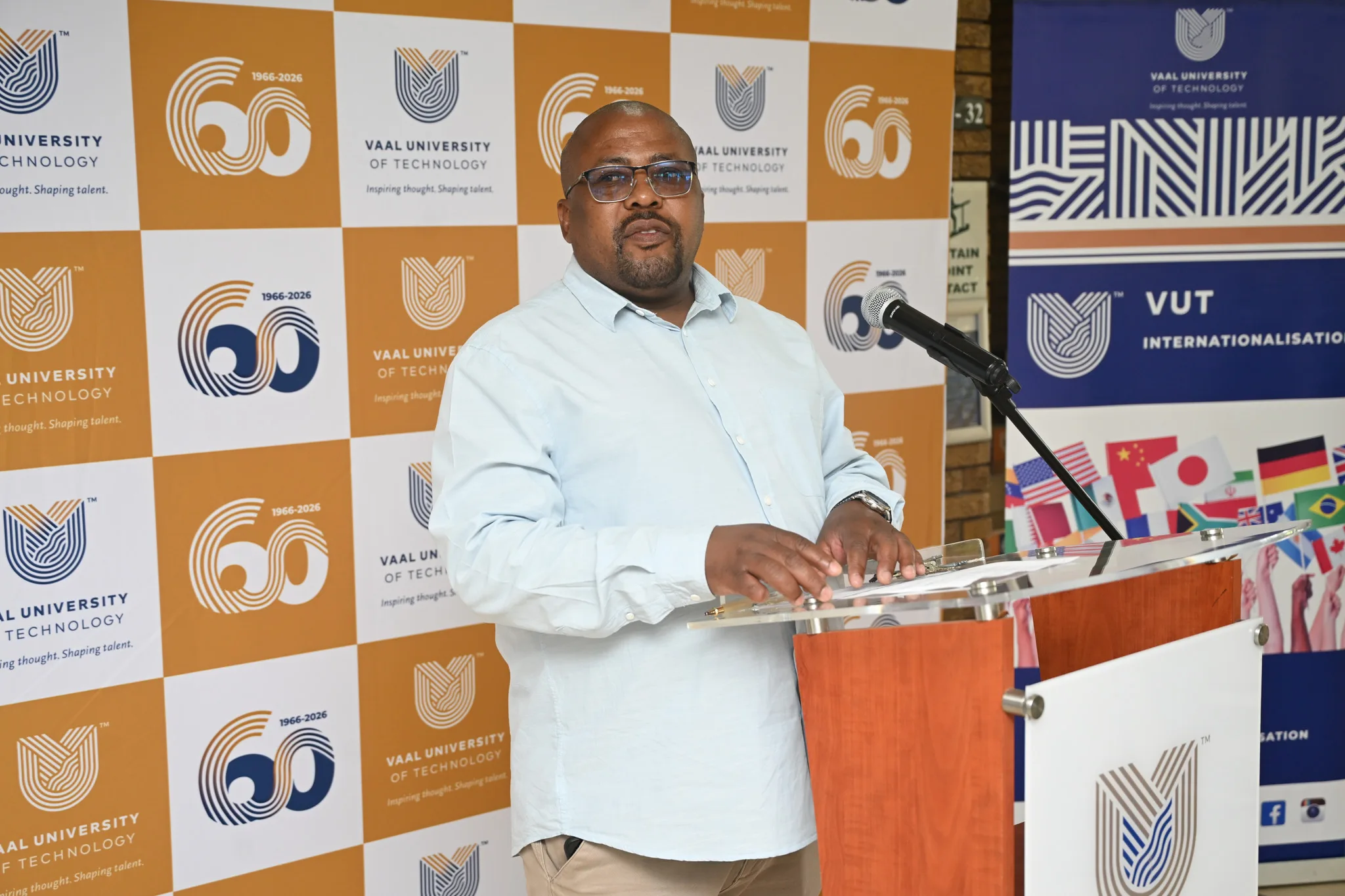 Man wearing glasses speaks at a podium with a microphone, in front of a checkered orange‑and‑white backdrop with Vaal University of Technology logos.