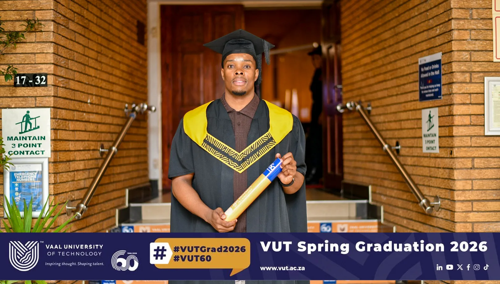 PRUDYKAY_VIEWS – Vaal University of Technology – Vaal University of Technology Graduate in cap and gown holding a diploma outside a brick building with a VUT Spring Graduation banner in the foreground