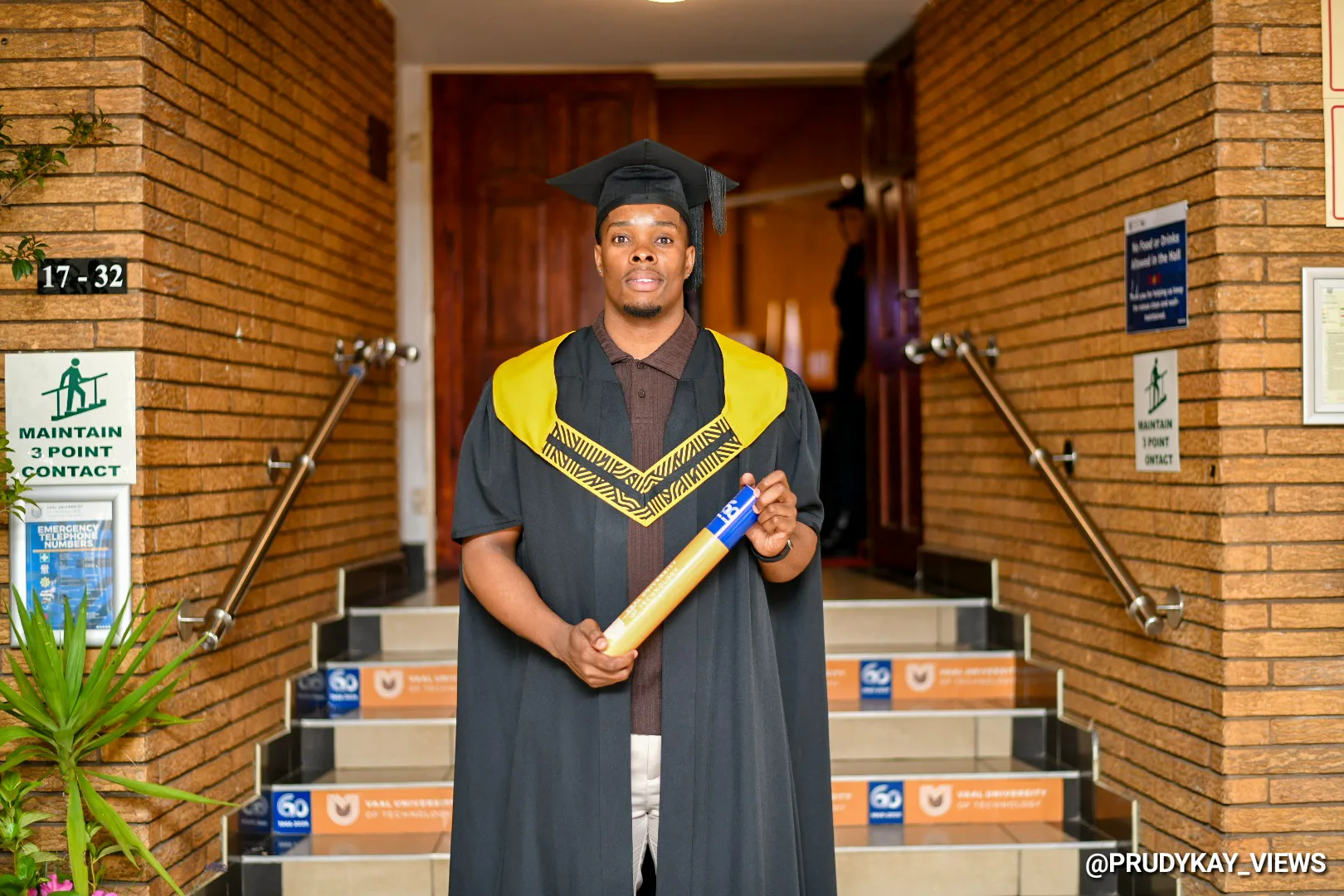 Graduate in cap and gown holding a diploma outside a brick building, standing on steps under a doorway.