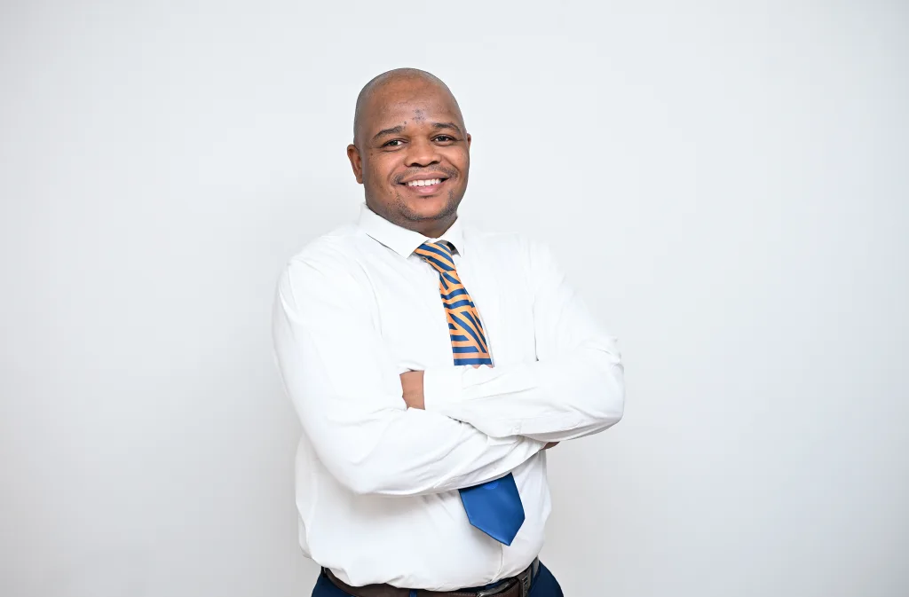 Smiling man in a white shirt and orange-blue striped tie stands with arms crossed against a light gray background, portrait style.