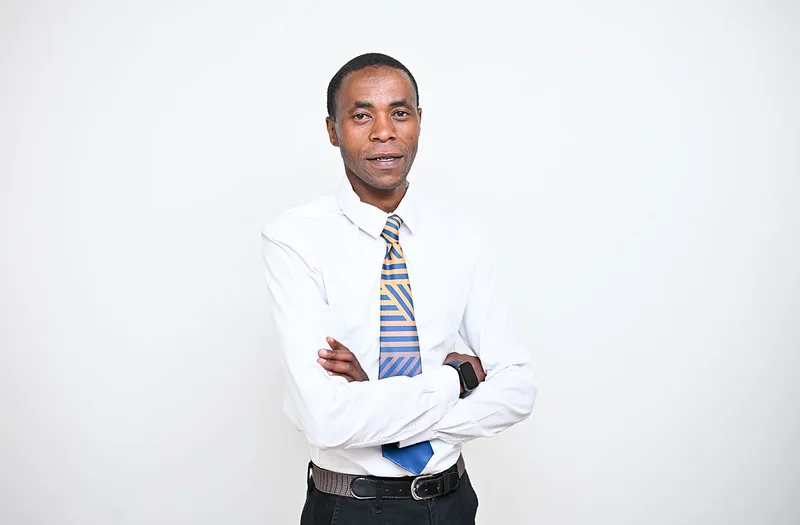 Professional man in a white dress shirt and striped blue–gold tie stands with arms crossed against a pale background.