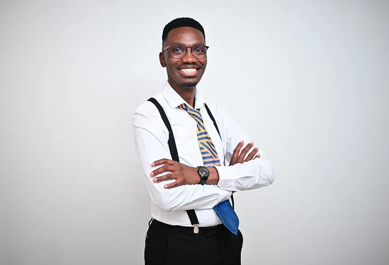 Smiling Black man in a white dress shirt with suspenders and a striped tie, arms crossed, standing against a light background.