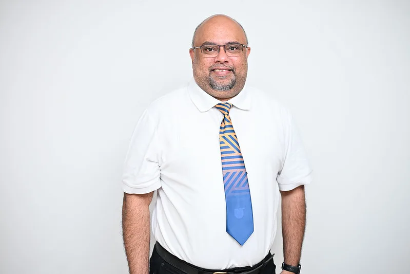 Smiling bald man with glasses in a white shirt and blue-yellow patterned tie, posing for a portrait against a light background