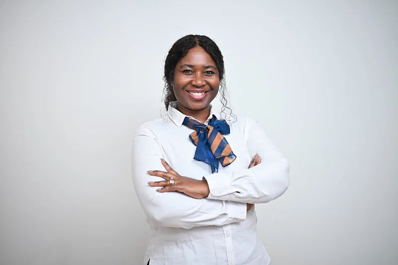 Confident woman with arms crossed, wearing a white blouse and blue scarf, smiling against a light gray background.