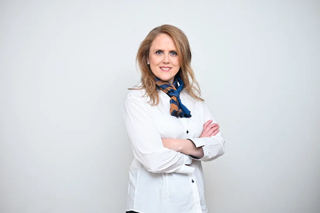 Professional woman with hands folded, wearing a white shirt and blue scarf, smiling against a light gray background, looking at the camera.
