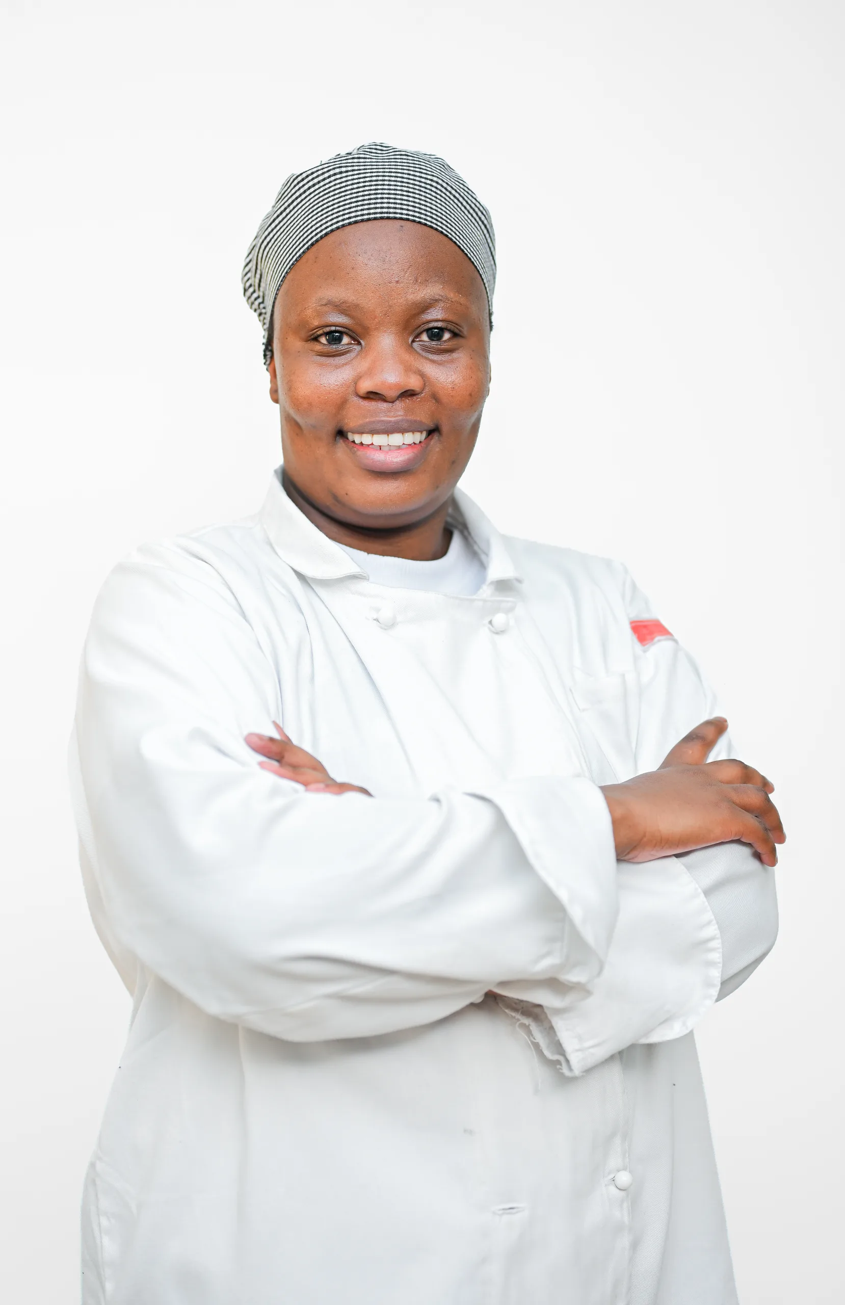 Smiling Black woman in a white lab coat with a striped head wrap, arms crossed against a light background.