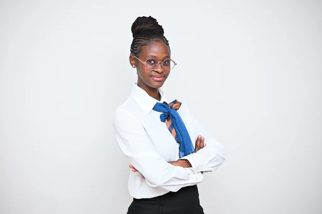 Professional young woman with braided hair and round pink glasses, wearing a white blouse and blue scarf, arms crossed against a light gray background.