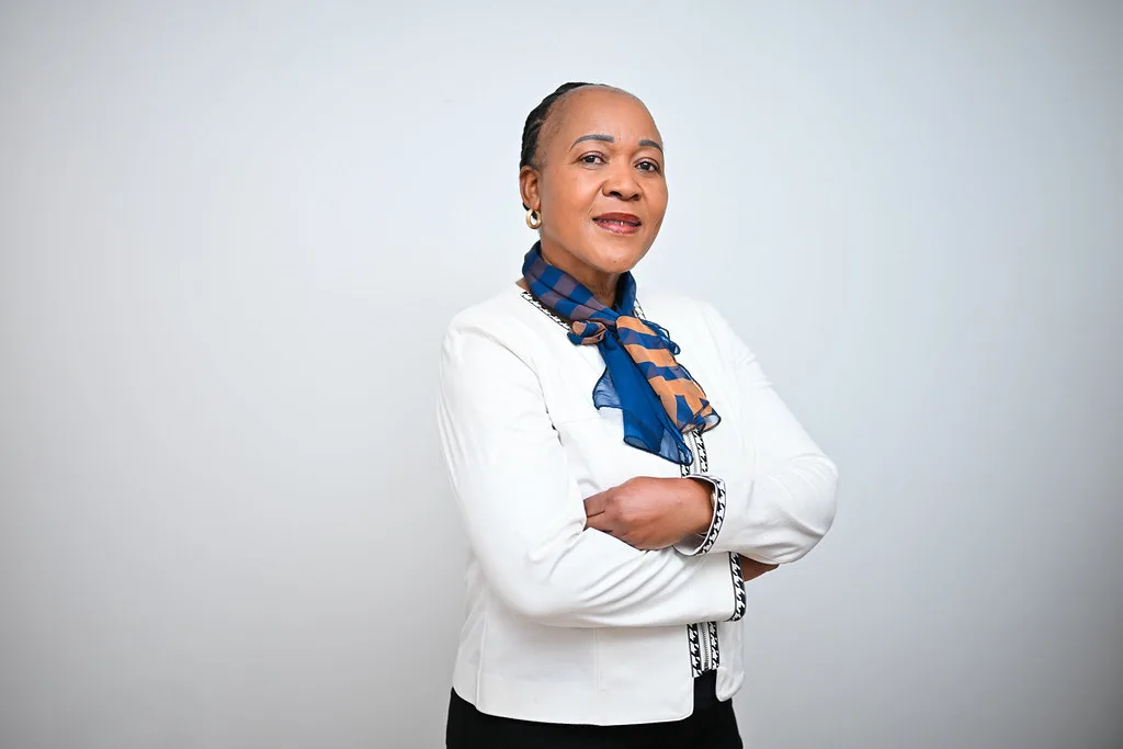 Confident woman with arms crossed wearing a white blazer and blue scarf against a light background.