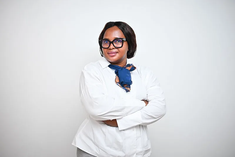 Professional woman with dark hair and glasses, wearing a white blouse and blue scarf, arms crossed, posing confidently against a light background.