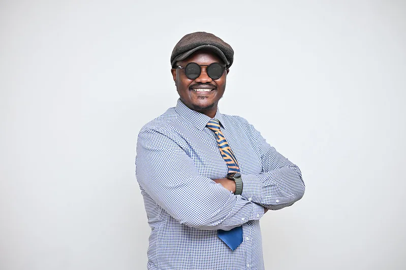 Smiling man in a flat cap and round sunglasses, wearing a blue checkered shirt and striped tie, arms crossed.