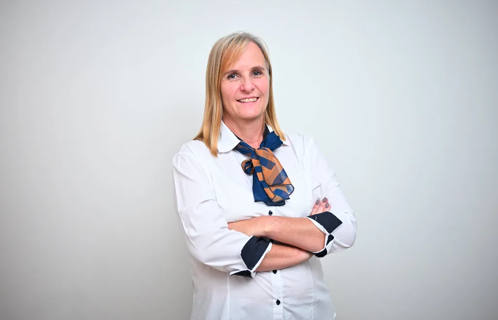 Professional woman with blonde hair in a white blouse and checkered scarf, arms crossed, smiling against a neutral background.