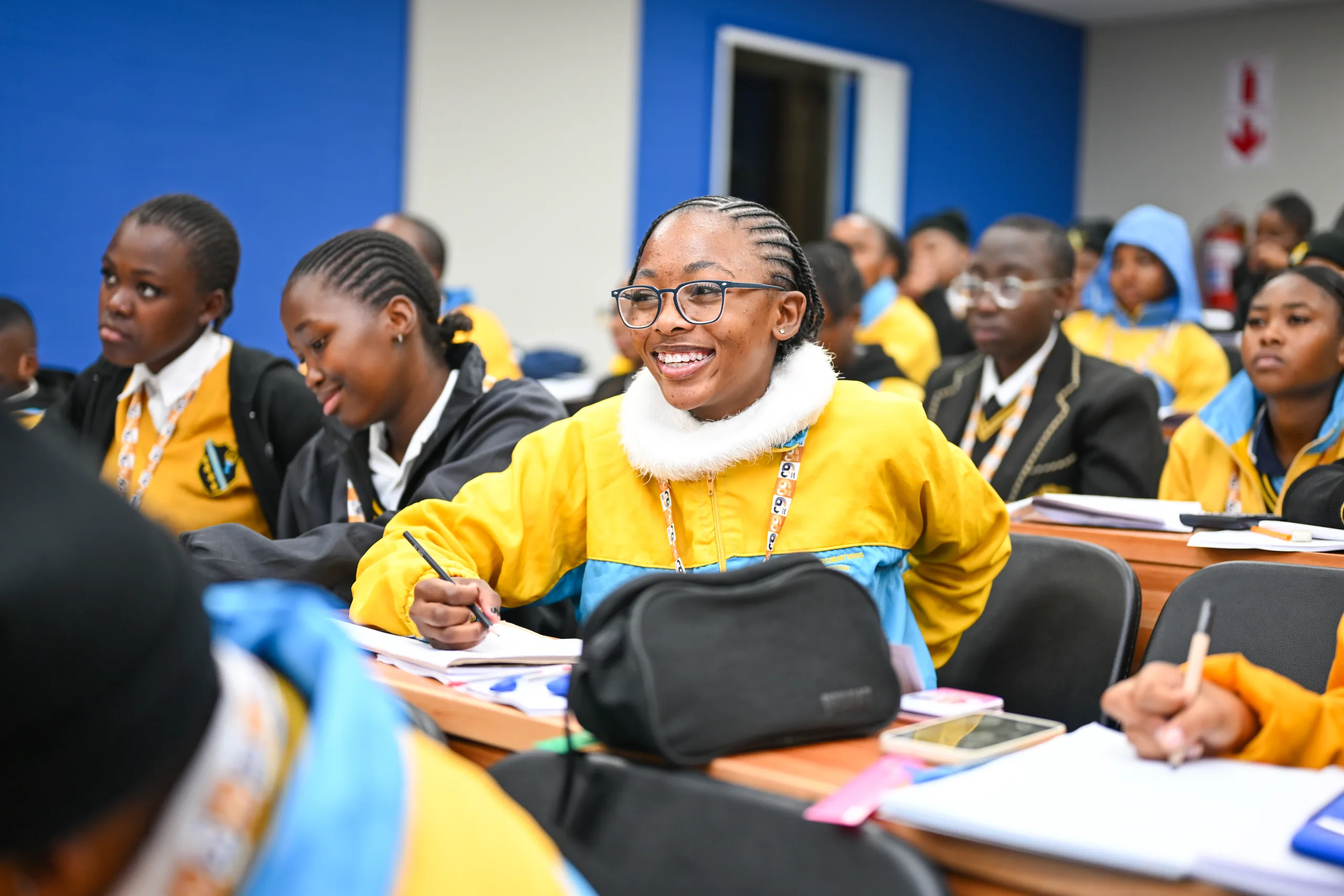 VUT and LGSETA – Vaal University of Technology – Vaal University of Technology Smiling student in yellow and blue uniform writes in a notebook in a classroom full of peers. central figure wears glasses and braided hair.